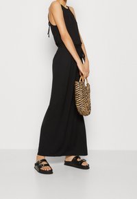 Svart ärmlös jumpsuit med knytmidja, kombinerad med svarta platta sandaler och en vävd brun handväska med cirkulär design och texturerad yta.