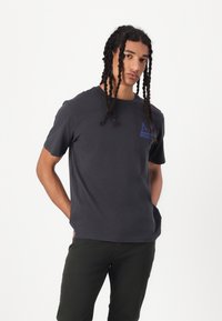 Camiseta de algodón gris con cuello redondo y mangas cortas, que presenta un logo azul en el lado izquierdo del pecho, combinada con pantalones verde oscuro.