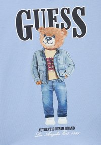 Camiseta azul claro con un oso que lleva una chaqueta vaquera, camiseta gráfica y vaqueros. El texto incluye "GUESS" y "AUTHENTIC DENIM BRAND".