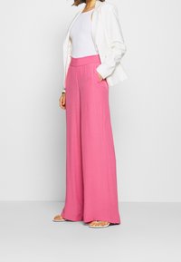 Pantalons larges roses en tissu doux, avec une taille haute et des poches, associés à un blazer blanc et un débardeur ajusté.