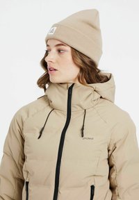 Beige Daunenjacke mit hohem Kragen, schwarzem Reißverschluss und Kordeln. Getragen mit einer hellbeigen Mütze. Dezente Markenkennung auf der Jacke sichtbar.