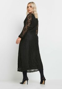 Svart spets maxiklänning med långa ärmar, genomskinliga detaljer på armarna och en flödande silhuett. I kombination med svarta högklackade skor och strumpbyxor.