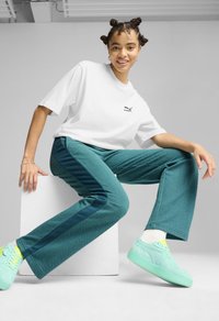 Bela majica s kratkimi rokavi s Puma logotipom, teal hrapave hlače s temno zelenimi stranicami, meta zelene športne čevlje, sedenje na belem podstavku.