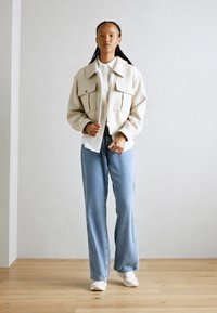 Beige oversized jakke med to brystlommer, lagd over en hvit skjorte, sammen med lyseblå vide jeans og hvite sneakers.