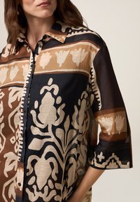 Eine knöpfbare Bluse mit einem floralen und geometrischen Muster in Braun, Creme und Marineblau. Hergestellt aus leichtem Stoff mit einer lockeren Passform und kurzen Ärmeln.