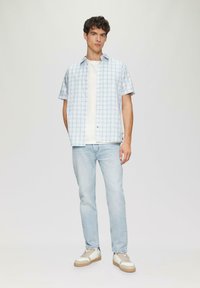 Korte mouwen, lichtblauw geruit shirt met witte accenten, gedragen over een wit t-shirt. Gecombineerd met lichtblauwe jeans en beige sneakers.