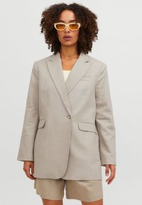 Beige zakó egysoros gombolással, notch gallérral, két elülső zsebbel és laza fazonnal, bézs sorttal és sárga napszemüveggel párosítva.