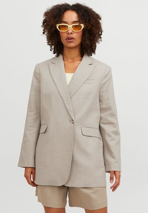 Blazer beige avec une fermeture à un bouton, revers crantés, deux poches avant et une coupe décontractée, associé à un short beige et des lunettes de soleil jaunes.
