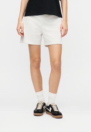 Person trägt hellgraue Freizeithose, schwarze Sneakers mit weißen Details, weiße Socken und ein schwarzes Oberteil, steht vor einem weißen Hintergrund.