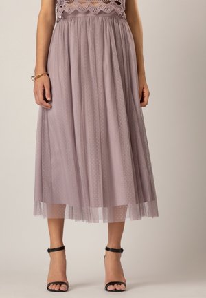 Femme portant une jupe midi en tulle mauve à pois et un haut en dentelle, associée à des sandales à talons hauts noires avec bride à la cheville.