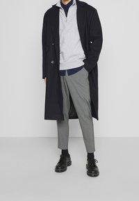 Abrigo largo azul marino sobre un sudadera gris con cremallera, superpuesta a una camisa azul, combinada con pantalones grises ajustados y zapatos negros con cordones.