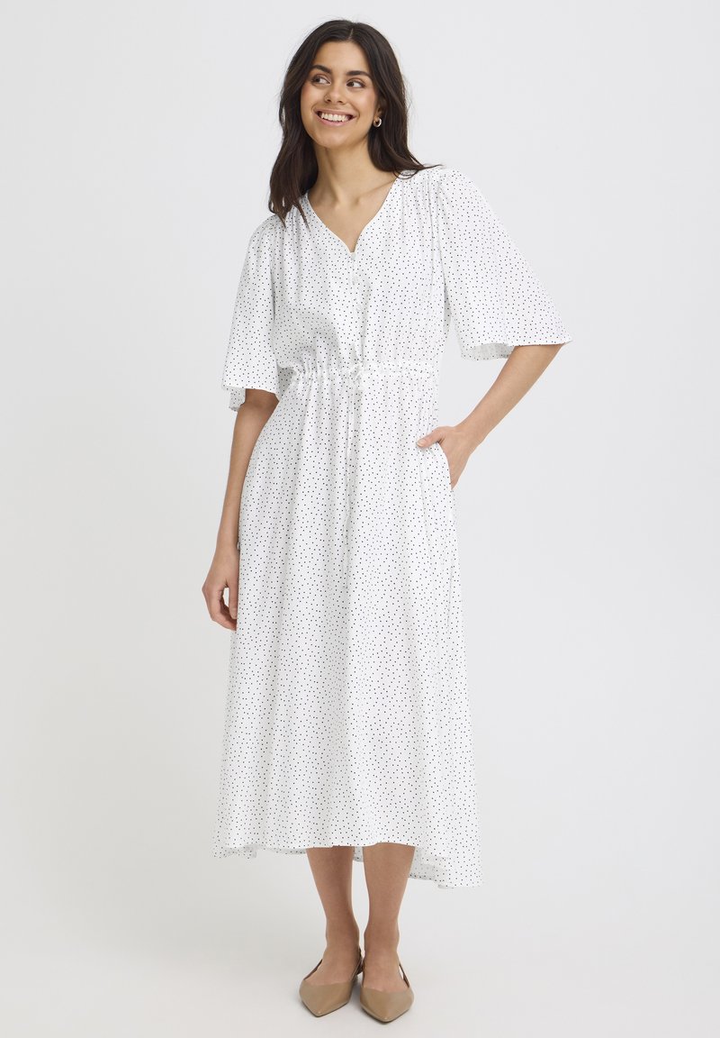 Femme souriante, portant une robe midi blanche à pois noirs, manches courtes larges, taille cintrée, et des ballerines beiges pointues, debout sur un fond uni.
