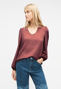 Γυναίκα με ξανθά μαλλιά που φοράει μια ροζ μπλούζα με υφή και μακριά μανίκια, καθώς και μπλε ψηλόμεσα τζιν με διακοσμητικά σχέδια κοντά στα γόνατα.