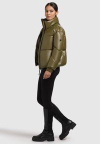 Olive green puffer jacket with a high collar, quilted design, and zip closure. Worn over black leggings and ankle boots with chunky soles.