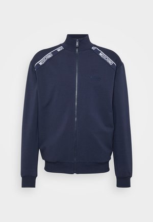 Jachetă de marină cu fermoar, guler înalt, manșete și tiv cu model în relief. Prezintă benzi albe cu marcaj pe umeri și un material texturat.