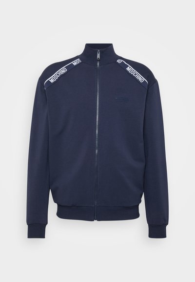 Jaqueta de fecho éclair azul-marinho com colarinho alto, punhos e bainha canelados. Apresenta fita branca com marca nos ombros e tecido texturizado.