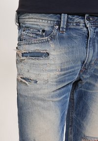 Ljusblå denimjeans med slitna områden, synlig sömnad och en blekt textur. Inkluderar flera fickor, med fransade kanter.