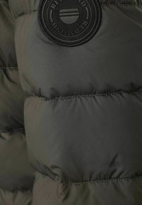 Kurtka puchowa w oliwkowym kolorze z pikowaną teksturą. Posiada czarną, okrągłą naszywkę z tłoczonym logo. Gładkie, błyszczące wykończenie.