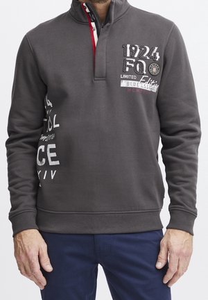 Mężczyzna ubrany w ciemnoszary długi sweter z napisem „1924 FQ Limited Edition” oraz granatowe spodnie, stoi ze swobodnie opuszczonymi rękami.