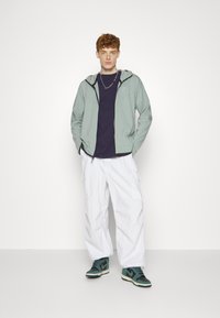 Lichte groene ritsjack over een donkerpaarse shirt, witte cargobroeken en groen/zwart sneakers. Eenvoudig ontwerp met een casual pasvorm.