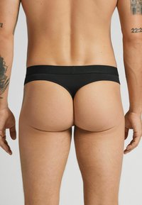 Schwarzse Thong-Unterwäsche aus glattem Stoff mit breitem Bund, entworfen um minimalen Bereich zu bedecken und eine klare, einfache Silhouette zu präsentieren.