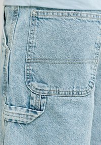 Poche de pantalon cargo en denim bleu clair avec rabat et détails cousus, portée sous une chemise ou un vêtement de couleur claire.