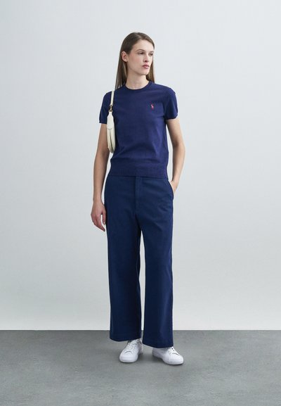 Μπλε σκούρο πουλόβερ με κοντά μανίκια και μικρό λογότυπο, σε συνδυασμό με φαρδιά μπλε σκούρα παντελόνια και λευκά αθλητικά παπούτσια, ολοκληρωμένο με μια λευκή τσάντα.