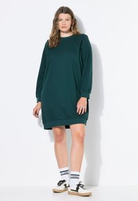 Dunkelgrünes Sweatshirt-Kleid mit langen Ärmeln, gerippten Details, rundem Halsauschnitt und lässiger Passform, kombiniert mit weißen Socken und schwarzen Turnschuhen.