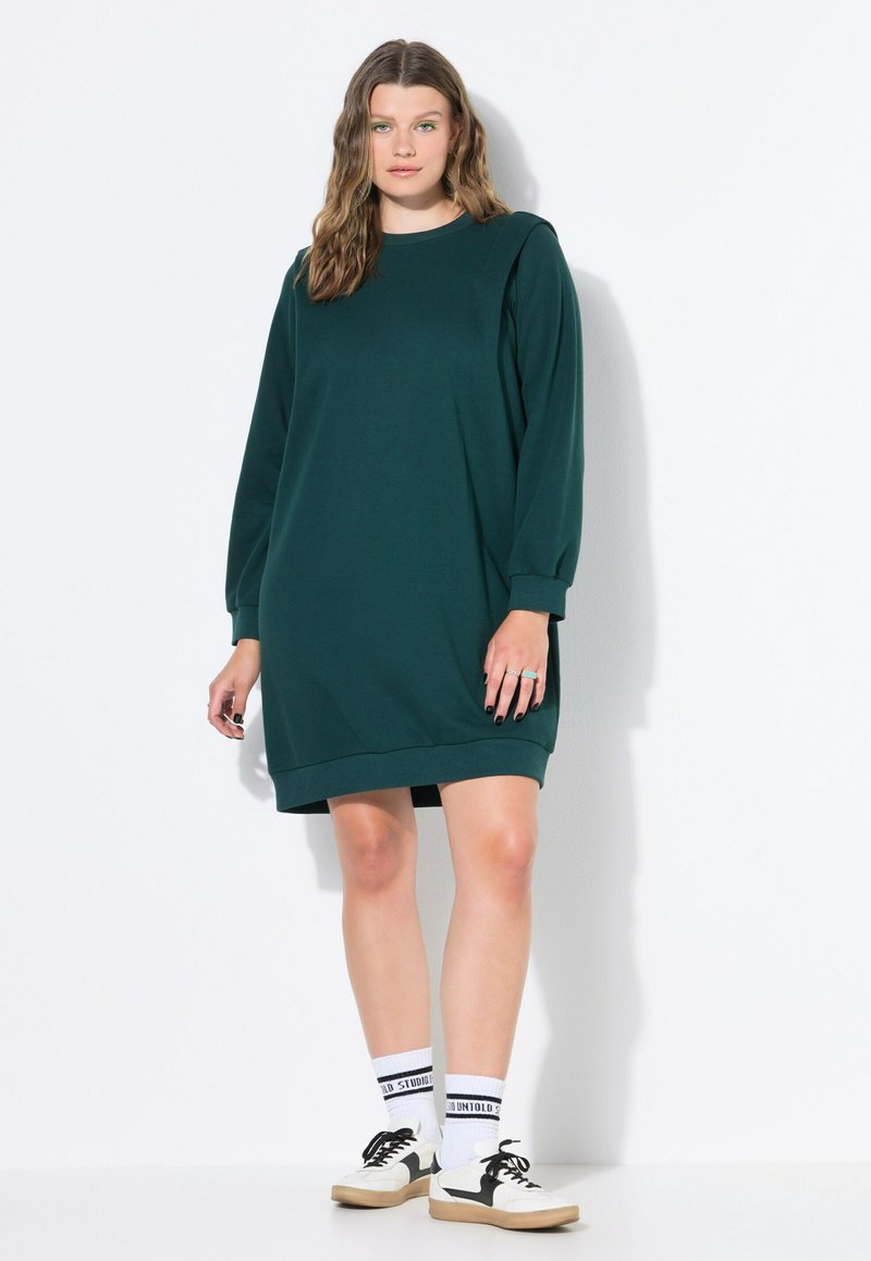Dunkelgrünes Sweatshirt-Kleid mit langen Ärmeln, gerippten Details, rundem Halsauschnitt und lässiger Passform, kombiniert mit weißen Socken und schwarzen Turnschuhen.