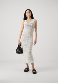 Robe maxi en crochet blanc avec des motifs géométriques, un décolleté en forme de goutte et une silhouette ajustée. Associée à des accessoires noirs et à des sandales.