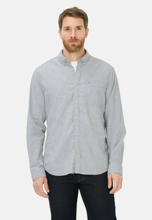 Homme aux cheveux bruns et barbe, portant une chemise à rayures gris clair boutonnée et un pantalon foncé, debout devant un fond blanc uni.