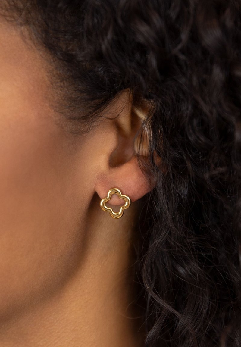 Goldene Ohrstecker in einem Vierblattmuster mit glatter Oberfläche und hohlem Zentrum. Tragen von einem Modell mit dunklem, lockigem Haar.