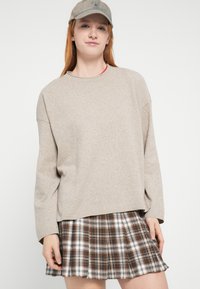 Μπεζ πλεκτό πουλόβερ με μακριά μανίκια και χαλαρή εφαρμογή, συνδυασμένο με καφέ και λευκή πλισέ φούστα με καρό σχέδιο. Συνοδεύεται από ξεθωριασμένο καπέλο.