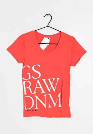 Rood T-shirt met korte mouwen en grote witte gedeeltelijke tekst "GS RAW DNM" op de voorkant gedrukt, hangend aan een zwarte hanger met witte labels.
