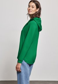 Zelená mikina s kapucí a dlouhými rukávy, zakončená zakřiveným lemem, spolu s světle modrými džíny. Tkanina vypadá hladce a ležérně.