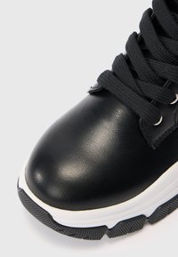 Adidași negri din piele cu talpă groasă texturată albă și neagră și șireturi negre legate într-o fundiță, pe un fundal simplu.