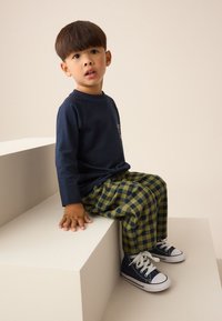 Temno modra majica z dolgimi rokavi, zelene in črne karo hlače, mornarsko modri športni copati z belimi vezalkami, otrok sedi na beli geometrični platformi.