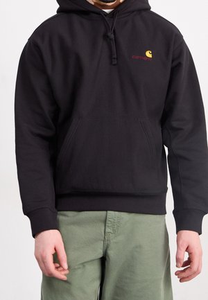 Mann trägt schwarzen Carhartt Hoodie mit Fronttasche und grünen Hosen, steht mit entspannten Armen seitlich am Körper vor weißem Hintergrund.