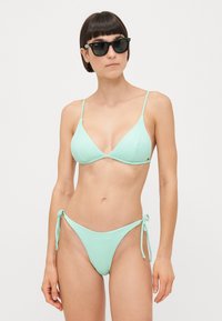 Myntegrønn bikini med ribbet tekstur, med tynne justerbare stropper og bunn med knytekanter. Brukt med svarte solbriller.