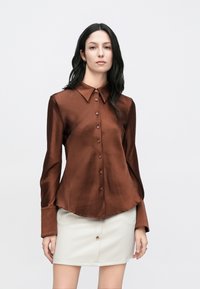 Braunes Satinkleid mit spitzem Kragen, Knopfleiste vorne und langen Ärmeln, kombiniert mit einem weißen Rock mit vorderer Knopfdetail.