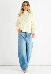 White knitted sweater with textured bobble details and ribbed collar, paired with light blue wide-leg jeans and pale pink flat shoes.