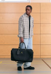Svart duffelbag med gullfargede detaljer, håndtak på toppen og skulderstropp, med teksturert stoff og en Jordan-logo. Modellen har på seg en rutete skjorte og jeans.