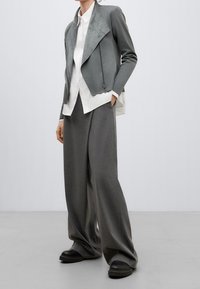 Giacca in ecopelle grigia con un ampio collo, abbinata a una camicia bianca e pantaloni larghi grigi. Le scarpe con tacco alto nere completano l'outfit.