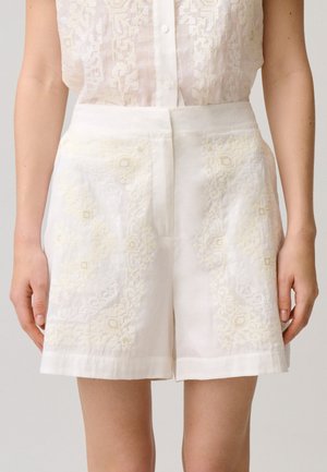 Shorts blancos de talle alto con sutiles patrones geométricos bordados, combinados con una blusa de encaje transparente abotonada.