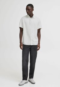 Camisa de botón de manga corta color gris claro con collar, combinada con jeans oscuros y zapatillas grises, de pie contra un fondo neutro.