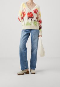 Pull en maille crème avec un col en V, orné de motifs floraux multicolores ; assorti à un jean bleu clair et des sabots gris à talons.