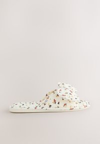 Chausson blanc matelassé à motif floral, à enfiler, avec un détail nœud sur le dessus, semelle plate et petite étiquette de marque en tissu sur le côté.