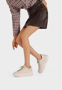 Donna che si inclina in avanti indossando una gonna in pelle marrone, una camicia a scacchi e sneakers beige con piattaforma e dettagli metallici su uno sfondo bianco.