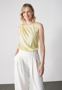Top senza maniche in satin giallo chiaro con frontale drappeggiato, abbinato a pantaloni bianchi a vita alta con pieghe, su uno sfondo neutro.