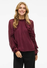Burgundy bluse med volumiøse detaljer og lange, transparente ærmer. Har en høj halsudskæring og en afslappet pasform, parret med sorte bukser.